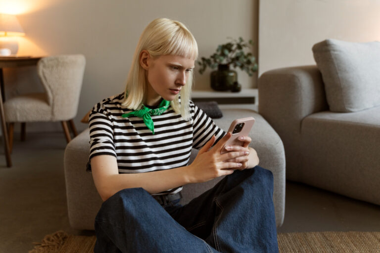 Žmogus naudojasi išmaniuoju telefonu, fone matomi psichikos sveikatos simboliai ir emocijų piktogramos